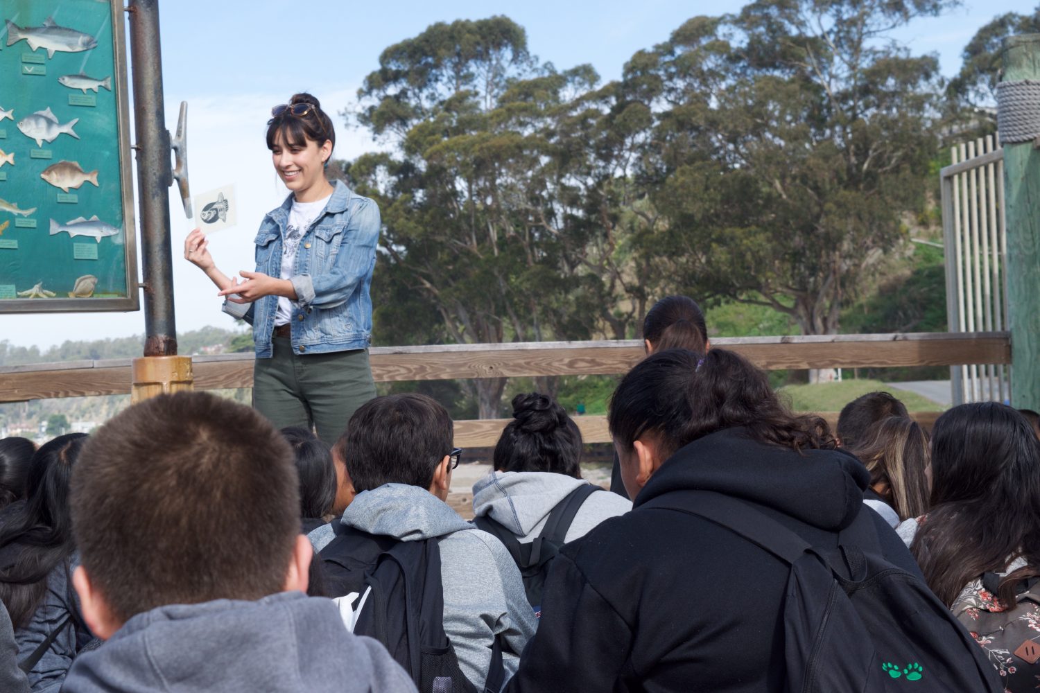 A woman with black hair holds a drawing and is showing it to a group of children that are sitting in front of her. They are all outside with big trees n the distance and a fence behind them.